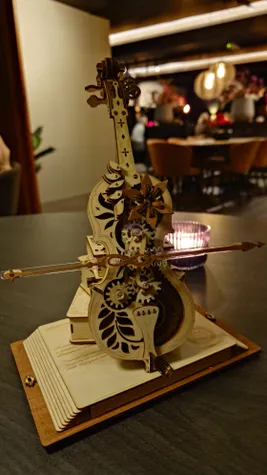 Een klein houten model van een cello op een opengeslagen boek met achter de cello wat andere boeken, de cello is aangedreven door tandwielen en maakt muziek zonder een bespeler hier van schuinvoor genomen