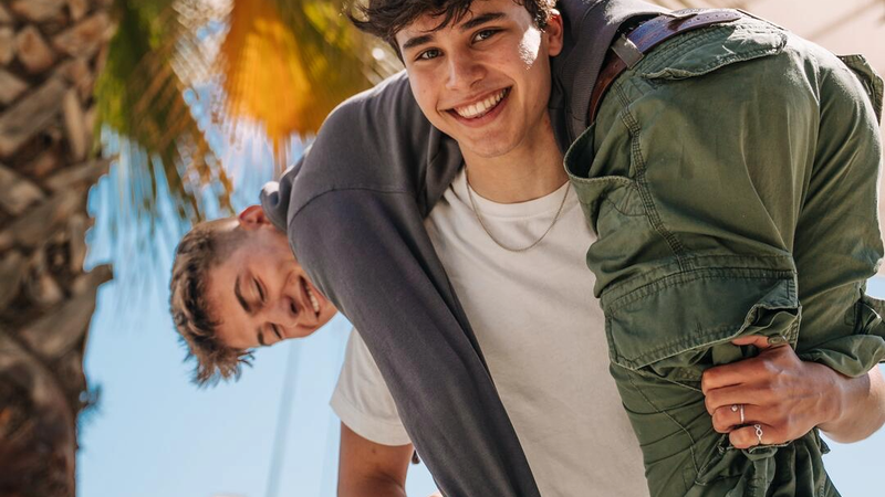Two EF Language Abroad students having fun outdoors in Málaga, Spain, with one playfully carrying the other over his shoulder in a sunny waterfront area lined with palm trees.