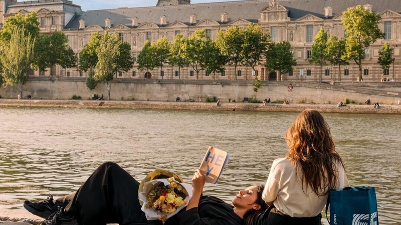 Two EF Language Abroad students relaxing by the Seine in Paris, reading and enjoying the riverside view across from the Louvre on a sunny afternoon.