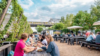 EF Language Abroad students eating and talking together at an outdoor beer garden in Berlin, with long picnic tables, greenery, and an elevated yellow U-Bahn train passing in the background.