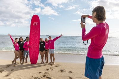 EF Language Abroad students posing with a bright pink surfboard on the beach in Costa Rica, smiling as a friend takes their photo by the ocean.