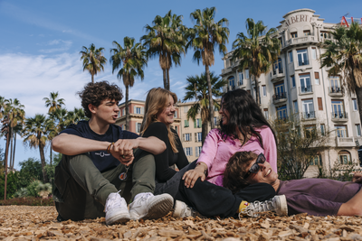 EF Language Abroad students relaxing and laughing together in a sunny park in Nice, France, sitting on the ground under palm trees with historic buildings in the background.
