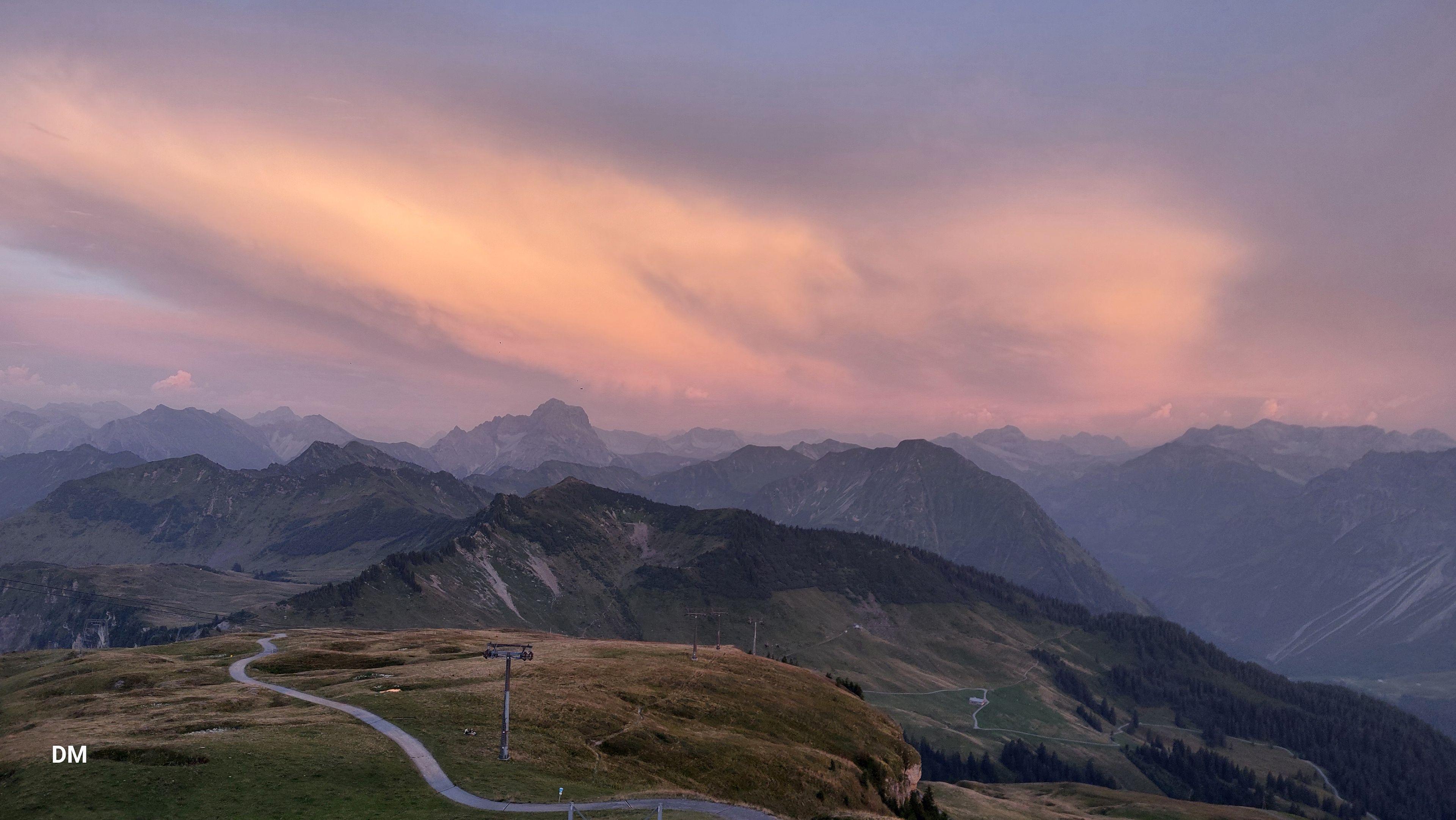 Berglandschaft bei Sonnenuntergang, mit pastellorangen Wolken und gewundenen Pfaden durch grüne Hügel, vor dem Hintergrund entfernter Gipfel.