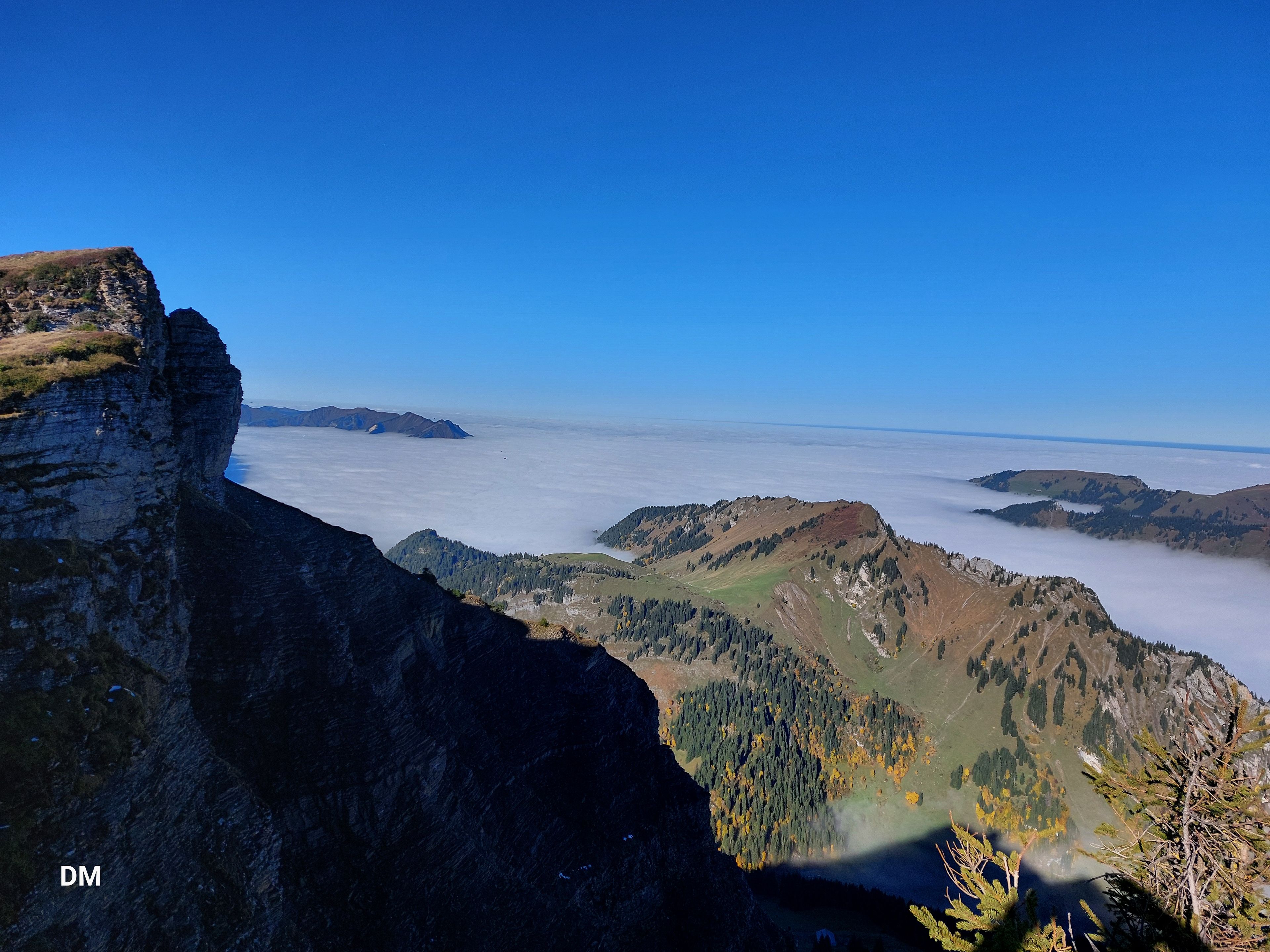 Berglandschaft mit schroffen Klippen auf der linken Seite, bewaldeten Hängen darunter und einem Meer aus Wolken, das sich unter einem klaren blauen Himmel bis zum Horizont erstreckt.