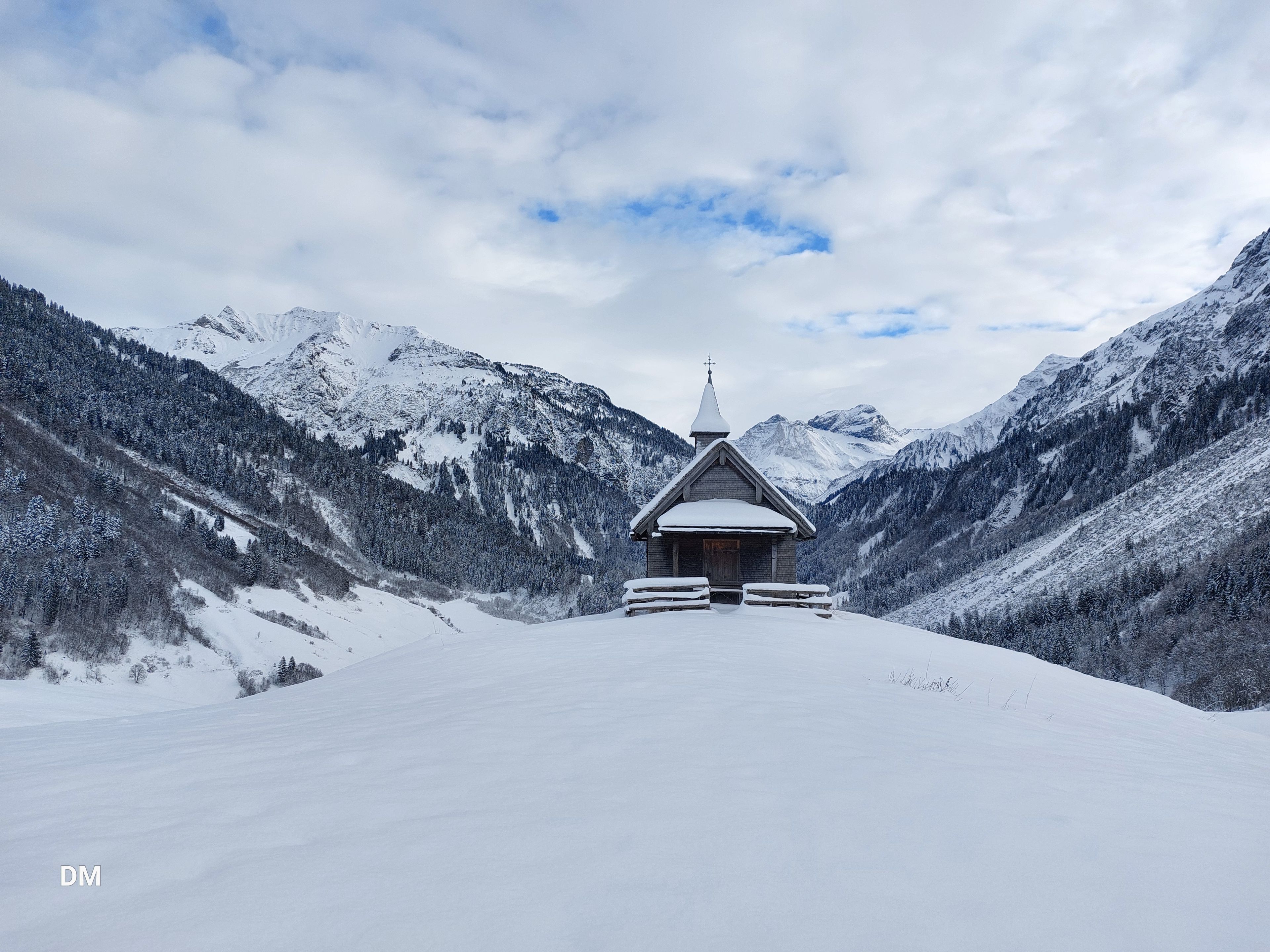 Eine kleine Holzkapelle steht auf einem verschneiten Hügel, umgeben von schneebedeckten Bergen und einem bewölkten Himmel.