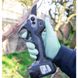 Person wearing green gloves using an electric pruning shear to cut a tree branch outdoors.