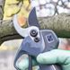 Gardener wearing green gloves uses electric pruners to cut a tree branch, with gears and blade visible.