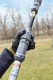 A person wearing a black glove holds a telescopic pole with warning labels in a grassy outdoor area.