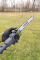 A gloved hand holding a small chainsaw with "ausonia" branding, set against a grassy outdoor backdrop with bare trees.