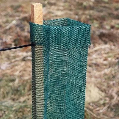 Green mesh fencing attached to a wooden stake, set against a blurred outdoor background with grass and soil.
