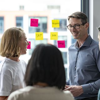 Four people discussing in front of a window covered with colorful sticky notes labeled with terms like 'SAP', 'MM', and 'Challenge', with buildings visible in the background.