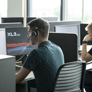 Two people working at desks in an office; one wears a headset and views a screen displaying 'XL2'.