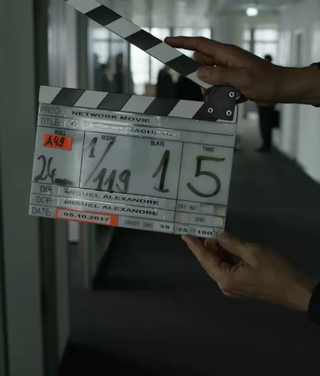 Hand hält weiß-schwarze Filmklapper in Büroflur mit Szenen- und Aufnahmeinformationen