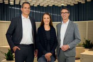 Drei Führungskräfte von XL2 stehen nebeneinander in einem modernen Büro mit blauem Vorhang im Hintergrund