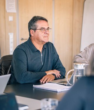 Mann mit Brille in blauem Poloshirt sitzt nachdenklich an Konferenztisch mit Laptop und Notizen