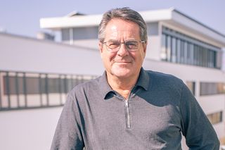 Mann mit Brille in grauem Polo-Shirt auf Bürogebäude-Terrasse, modernes Architektur-Setting im Hintergrund