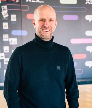 Mann mit Brille im blauen Hemd vor modernem Tech-Büro-Hintergrund mit Pflanzen und digitalen Designs