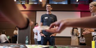Teilnehmer bei einem interaktiven Workshop-Spiel mit bunten Karten und Spielsteinen auf einem Tisch, während Felix Spitznagel im XL2-Shirt lächelnd moderiert