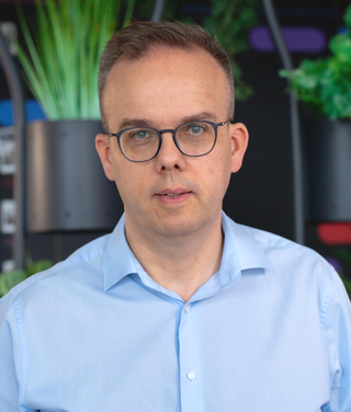Mann mit Brille im blauen Hemd vor modernem Tech-Büro-Hintergrund mit Pflanzen und digitalen Designs