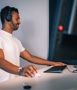 Mann am Schreibtisch mit Headset, bedient Computermaus vor Monitorwand in moderner Homeoffice-Umgebung