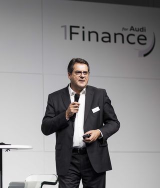 Geschäftsmann in schwarzem Anzug präsentiert mit Mikrofon vor Audi-Finance-Hintergrund auf Konferenz