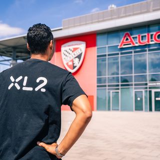 Mann von hinten im XL2-Shirt mit Fußball vor Audi Sport Gebäude
