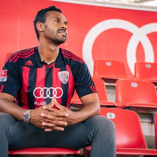 Mann in rot-schwarzem Fußball-Trikot sitzt nachdenklich auf roter Spielerbank im Stadion