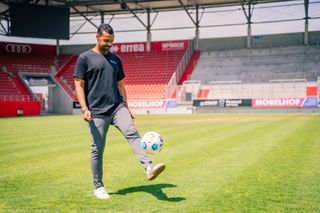 Mann in rot-schwarzem Fußball-Trikot sitzt nachdenklich auf roter Spielerbank im Stadion