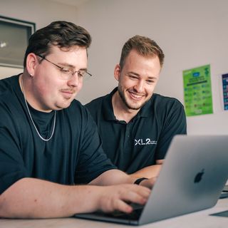 Zwei Mitarbeiter analysieren gemeinsam Daten auf einem Laptop vor großem Monitor