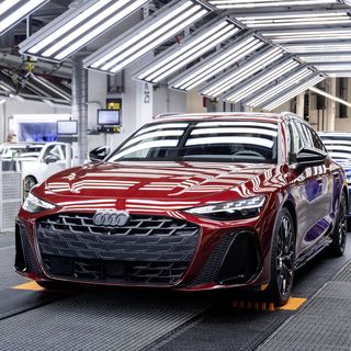 Red Audi car on a well-lit automotive production line, followed by other vehicles and surrounded by factory equipment.