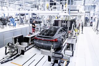 A grey Audi on an assembly line in a modern automotive manufacturing facility, surrounded by robotic arms and machinery. A worker in red shorts and a black shirt stands next to the car, monitoring the process.