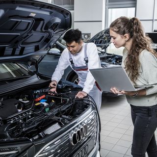 Two people working on an Audi with the hood open in a workshop; one inspects the engine while the other observes with a laptop.