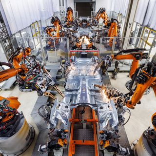 Luftaufnahme einer Fertigungshalle mit orangefarbenen KUKA-Industrierobotern beim synchronen Schweißen einer Fahrzeugkarosserie