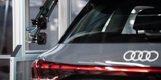 Rear view of a grey Audi car with a robotic inspection arm in an industrial setting.