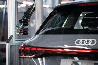 Rear view of a grey Audi car with a robotic inspection arm in an industrial setting.