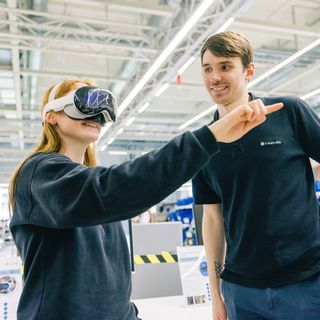 Zwei Mitarbeiter mit VR-Headset in modernem Büro mit weißer Industriedecke, einer zeigt begeistert etwas.