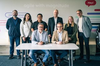 Achtköpfiges Team posiert professionell vor einer weißen Wand mit "agile with magnitude" Schriftzug