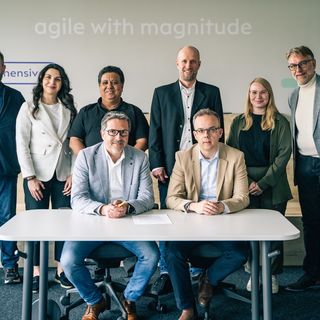 Achtköpfiges Team posiert professionell vor einer weißen Wand mit "agile with magnitude" Schriftzug