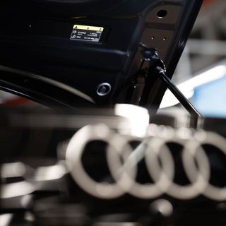 Open hood of an Audi car showing the hydraulic strut and a label, with the Audi logo in the foreground and a workshop in the background.
