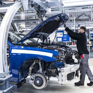Worker inspecting the engine of a blue car with the hood open in an automotive factory.