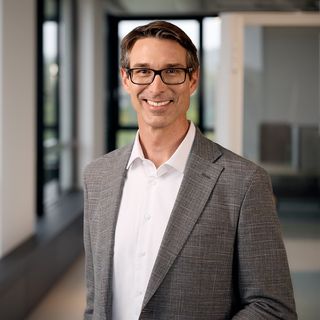 Lächelnder Felix Spitznagel mit Brille und grauem Sakko in modernem Büro mit Glaswänden