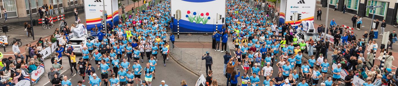 Hundreds of runners in blue shirts start the Wings for Life World Run in Breda under a large event arch surrounded by cheering crowds.