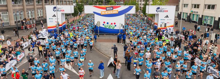 Hundreds of runners in blue shirts start the Wings for Life World Run in Breda under a large event arch surrounded by cheering crowds.