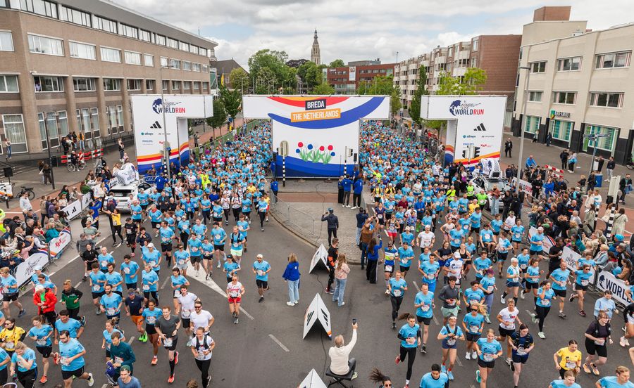 Hundreds of runners in blue shirts start the Wings for Life World Run in Breda under a large event arch surrounded by cheering crowds.