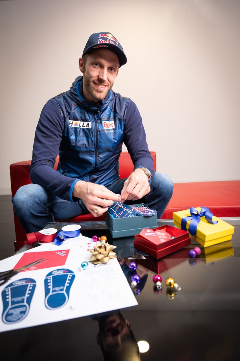 A man wearing a Red Bull cap and blue jacket sits on a red couch, wrapping gifts with ribbons and small ornaments on a table in front of him.