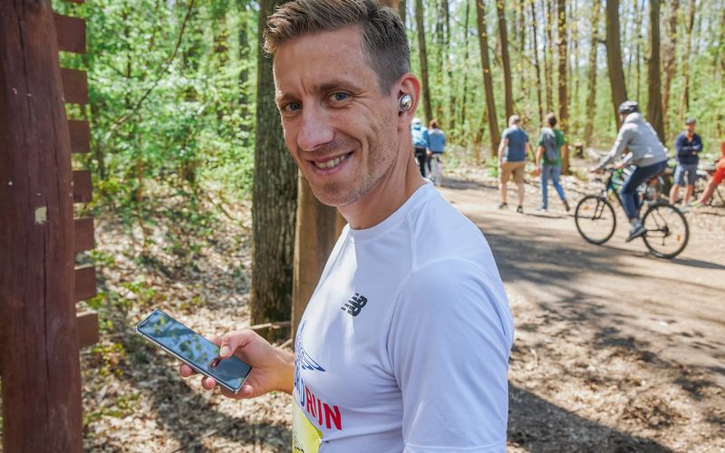 A man in a white Wings for Life World Run shirt smiles while holding a smartphone on a forest trail with people walking and cycling in the background.