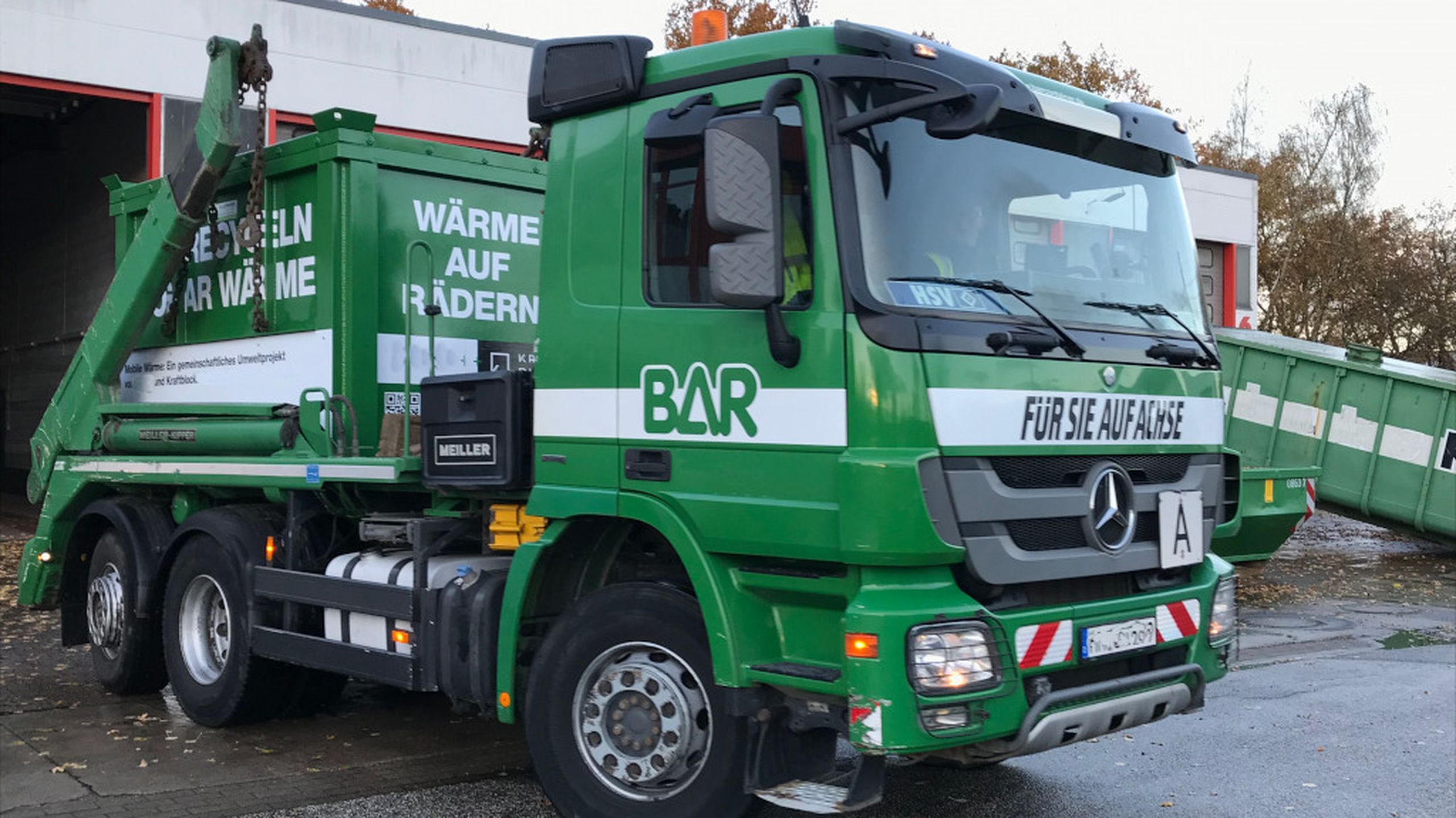 Green garbage truck parked outside a building, with messages and branding on the side. Trees and a clear sky in the background.