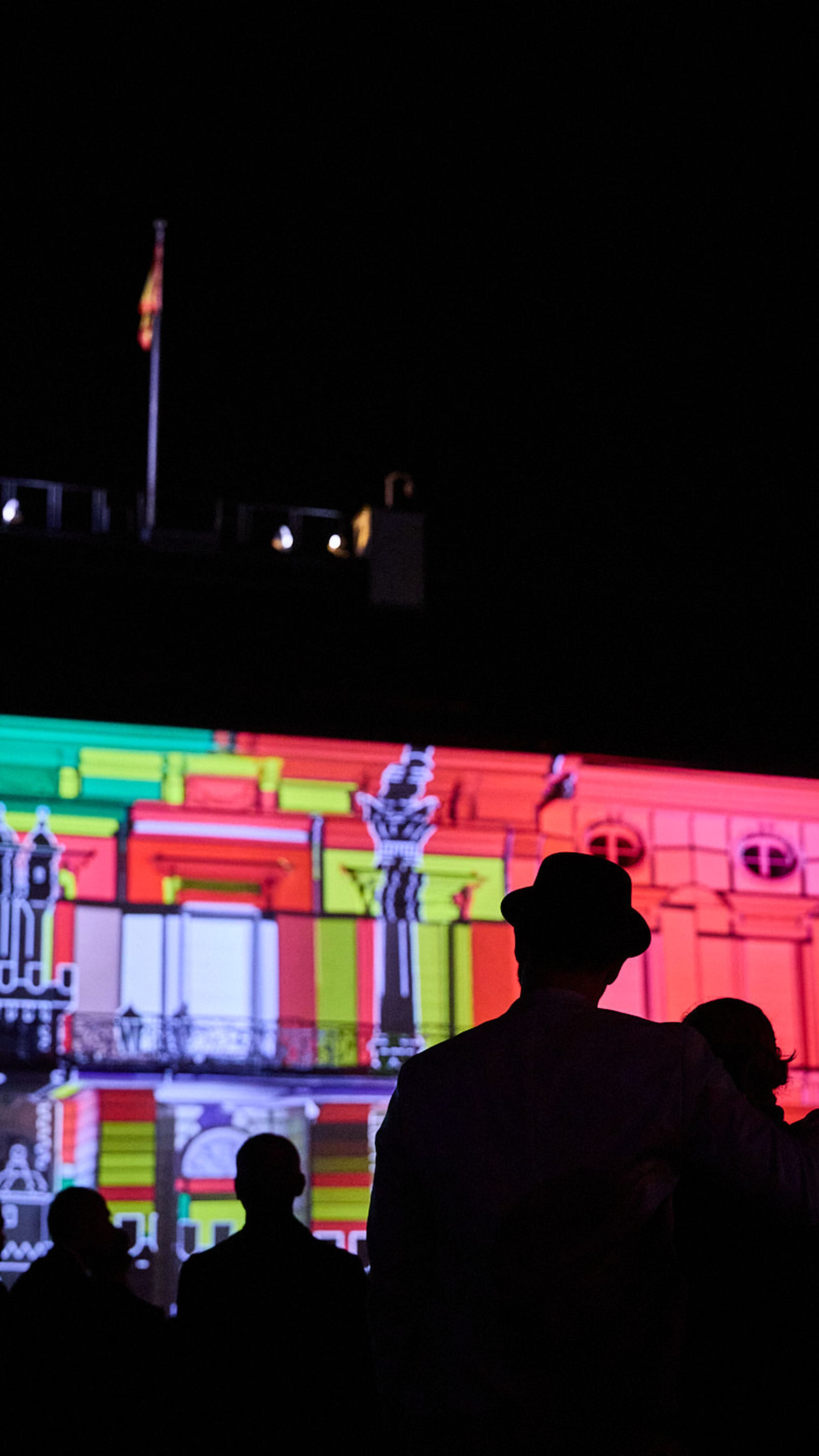Silhouetted crowd watches a colorful, illuminated building facade at night, featuring vibrant geometric and architectural projections.