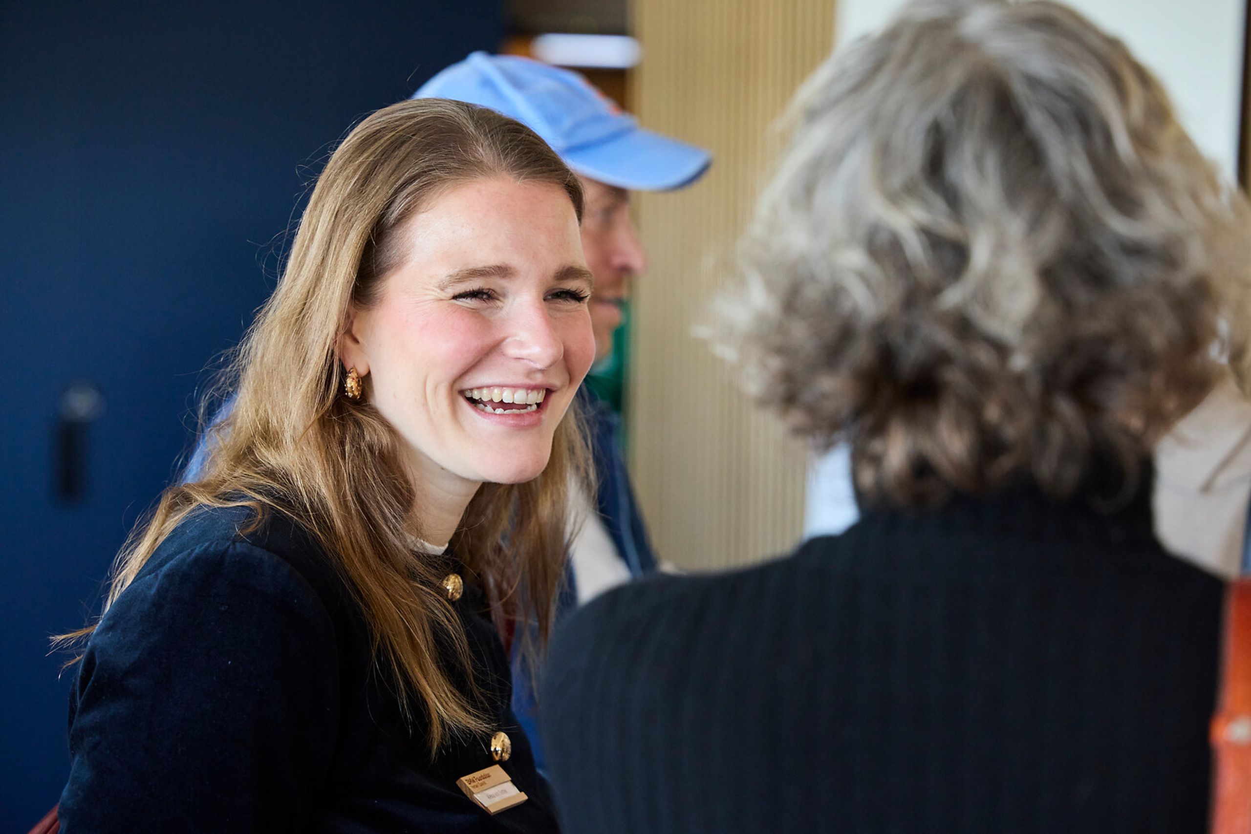 A woman with long hair smiles while talking to another person in a social setting. Other individuals are partially visible in the background.