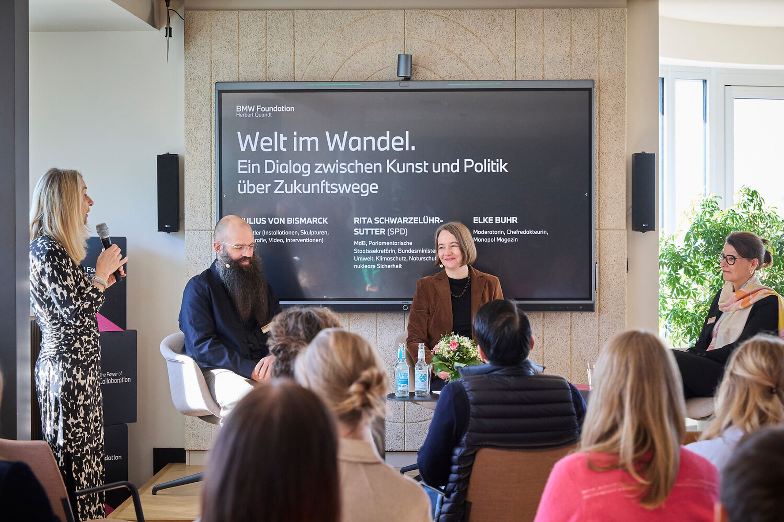 A panel discussion with three seated speakers and a standing moderator, with an audience in the foreground and a presentation screen behind them.
